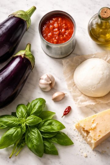Tous les ingrédients de la recette parmigiana d'aubergines disposés sur marbre : aubergines, mozzarella, parmesan, tomates concassées, basilic frais et huile d'olive