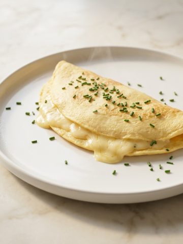 Omelette au fromage baveuse repliée sur assiette blanche avec ciboulette ciselée