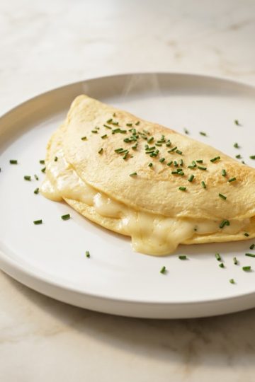 Omelette au fromage baveuse repliée sur assiette blanche avec ciboulette ciselée