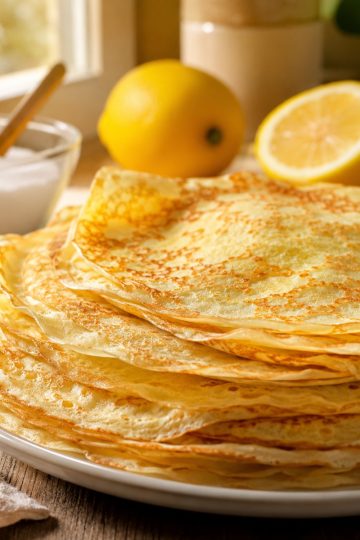 Pile de crêpes fines dorées maison sur une assiette, sucre et citron à côté